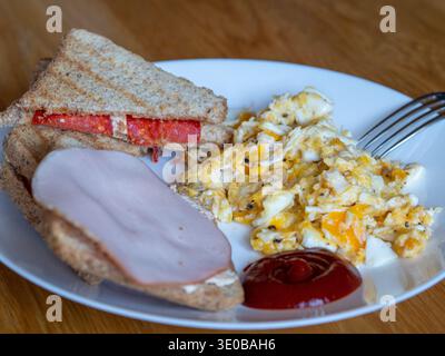 Sandwich appena tostato con prosciutto e pomodoro. Le uova strapazzate con erba cipollina si trovano accanto. Il ketchup rosso aggiunge un tocco di colore. Colazione semplice e salutare Foto Stock