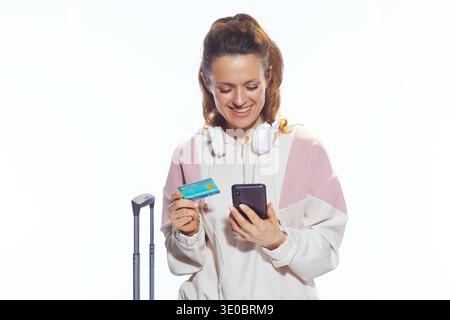 Una giovane donna sorride mentre usa il suo smartphone e la sua carta di credito per gestire i suoi piani di viaggio, stando accanto ai suoi bagagli in un luminoso studio. Foto Stock