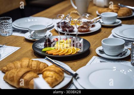 Primo piano di un tavolo da colazione con arance, uva e ananas tagliate a fette su un piatto nero, accompagnati da croissant freschi e marmellate. E Foto Stock