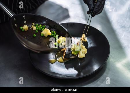 Primo piano di uno chef con guanti neri che prepara accuratamente verdure saltate su un piatto nero con pesce grigliato. Processo di placcatura culinaria di alta qualità in un p Foto Stock