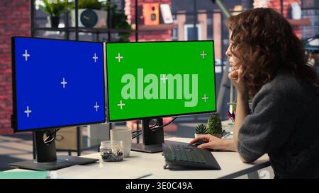 Dipendente in ufficio che lavora su un PC con spazio di copia isolato su due monitor, organizzazione creativa moderna. Donna concentrata su progettazione grafica, codifica o attività potenziate dall'intelligenza artificiale in uno spazio ottimizzato. Telecamera B.. Foto Stock