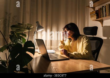 Donna stanca che soffre di ustioni e stress mentre lavora al suo notebook in un ambiente buio di ufficio domestico Foto Stock