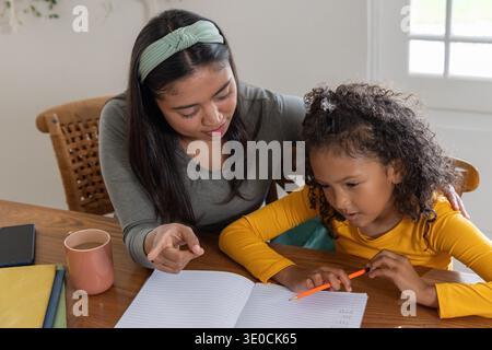 Il tutor guida il bambino, indicando il quaderno aperto sul tavolo della casa con una matita arancione e una tazza rosa Foto Stock