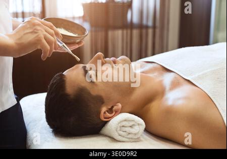 Estetista che applica lo scrub sul viso del giovane Foto Stock
