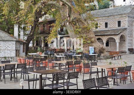 Tradizionale plateia greca del villaggio caratterizzata da un antico platano e tavoli da caffe' all'aperto vuoti Foto Stock