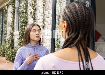 Due donne che indossano top atletici che praticano respirazione in uno stretto patio accanto a una porta scorrevole in vetro Foto Stock