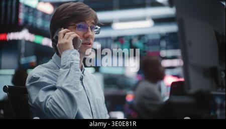 Broker di borsa focalizzato che gestisce i canali di comunicazione in Borsa. Man ha visto parlare attivamente sullo smartphone, confermare la transazione di valore elevato e monitorare gli indici digitali in background. Foto Stock