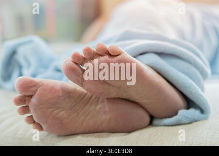 Primo piano di una donna anziana dorme a letto e coperta da una coperta blu. Concetto di assistenza ai piedi per anziani. Foto Stock