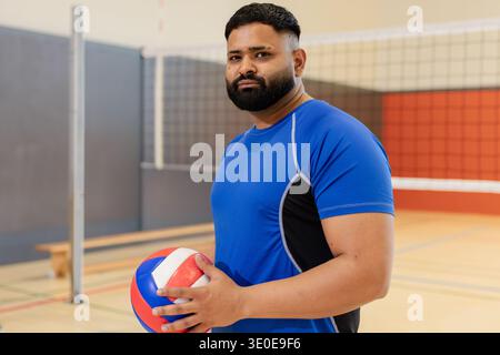 Uomo asiatico che tiene la pallavolo e sta in piedi sul campo da pallavolo vicino alla rete, indossando una maglietta blu atletica. Atleta, palla, palestra, panca, legno, vibrante, concentrato Foto Stock