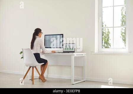 Analista donna che lavora al budget in ufficio domestico con monitor desktop e laptop Foto Stock