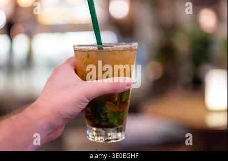Sorreggere a mano un bicchiere di mojito fresco con cubetti di ghiaccio e paglia verde. Drink estivo rinfrescante con rum e menta nell'atmosfera da bar, simbolo del nig Foto Stock