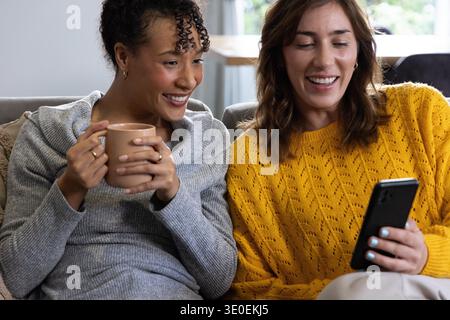 Diverse amiche si appoggiano insieme, condividendo lo smartphone sul divano nel soggiorno con una tazza beige. Duo, casual, accogliente, maglia, casa, mobile, illuminato dal sole || modello rilasciato Foto Stock
