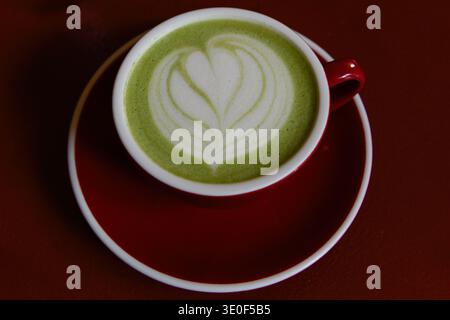 Tazza di tè matcha rosso e ceramica e piattino con cuore di latte in schiuma sulla superficie del tè. Il Matcha è una polvere finemente macinata di tè verde, specialmente proc Foto Stock