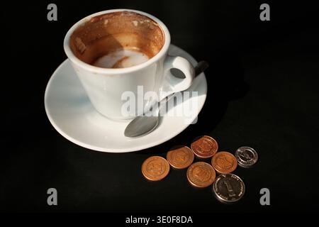 Cappuccino parzialmente bevuto in tazza bianca su piattino con monete posate sul tavolo. Concetto di pagamento del caffè, costo della pausa caffè e spese quotidiane in Foto Stock