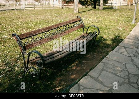 Vecchia panchina di legno con telaio metallico decorativo posta accanto a un vialetto di pietra nel giardino del parco. Concetto di relax, ricreazione all'aperto e pace Foto Stock