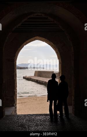 Due persone in sagoma guardano il mare attraverso l'antico arco in pietra di porta Pescara a Cefalù, in Sicilia. Lo sfondo mostra il molo e la spiaggia sabbiosa Foto Stock