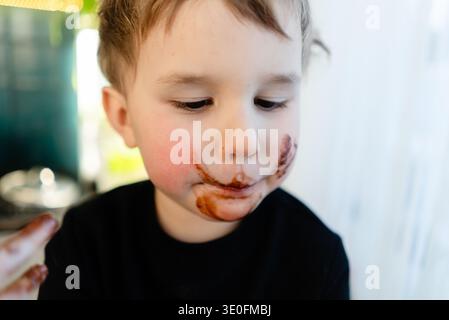 Ritratto di un bambino di tre anni con una faccia disordinata ricoperta di cioccolato. Contatto diretto con gli occhi dopo aver mangiato i dolci. Concetto di cattive abitudini alimentari infantili e conseguenze allergiche. Foto Stock