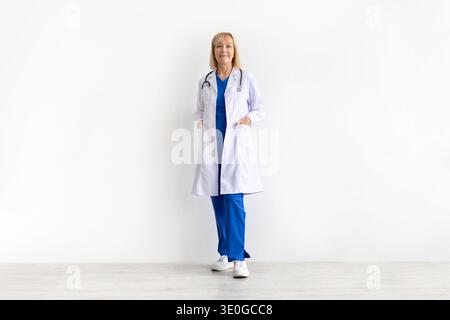 Medico donna anziana che indossa il cappotto medico bianco in piedi con sicurezza contro lo sfondo dello studio Foto Stock