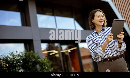 Una donna coreana sta fuori da un edificio moderno mentre usa un tablet. Sorride e sembra interagire con il dispositivo. L'impostazione è luminosa e. Foto Stock