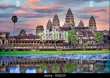 Il tempio di Angkor Wat si erge alto al tramonto. La gente del posto fa il bagno nello stagno di loto, riflettendo la bellezza di questo antico sito in Cambogia. Foto Stock