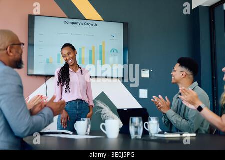 Giovane donna d'affari in piedi vicino allo schermo con grafico di crescita, mentre il team applaude nella sala riunioni dell'ufficio Foto Stock