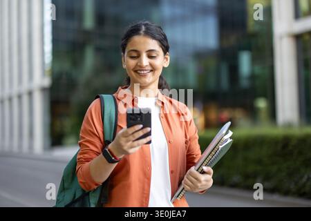 Giovane studentessa indiana sorridente con zaino e quaderni che controllano i contenuti sul suo cellulare mentre cammina all'aperto nel campus universitario, incarnando l'istruzione moderna e l'integrazione tecnologica Foto Stock