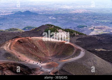 Vista aerea dei Crateri Silvestri sull'Etna. I turisti camminano lungo il bordo del cono vulcanico, caratterizzato da suolo rosso e nero. Sullo sfondo, Foto Stock