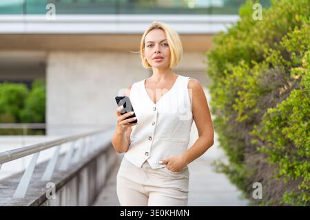 Giovane donna d'affari con i capelli biondi corti che indossa il gilet e i pantaloni che utilizzano lo smartphone in tutta sicurezza fuori dall'edificio dell'ufficio, accanto al verde Foto Stock