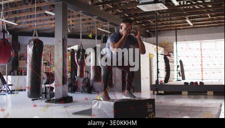 Uomo che atterra con abiti da allenamento scuri che esegue squat plyometrico su plyo box 20 nella palestra di boxe Foto Stock