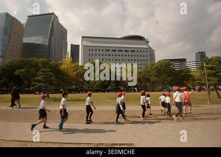 OSAKA, GIAPPONE - 14 NOVEMBRE 2025: Gli studenti delle scuole elementari giapponesi che indossano berretti rossi camminano in fila attraverso il parco del castello di Osaka durante una preparazione al disastro Foto Stock