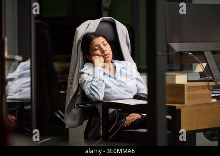 Giovane donna che dorme alla scrivania in un ufficio moderno, riposa a mano con gli occhi chiusi, indossa abiti da lavoro, siede sulla sedia da ufficio vicino al monitor del computer e ai documenti Foto Stock