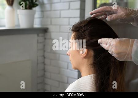 Le mani con i guanti tagliano con cura i lunghi capelli scuri delle donne. camera luminosa con trattamenti estetici per la cura dei capelli Foto Stock