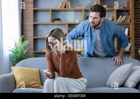 La donna si sente stressata e sconvolta, tenendo la testa mentre un uomo sta dietro il divano, esprimendo rabbia e gridando contro di lei durante un acceso litigio a casa Foto Stock