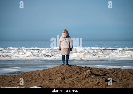 Una donna si trova su un'area sabbiosa vicino alla spiaggia che guarda all'oceano e al ghiaccio sull'acqua. Foto Stock