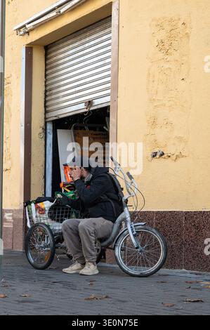Un uomo siede su un triciclo argentato con un cestello di filo metallico davanti a un magazzino di rottami metallici, in attesa di vendere i materiali raccolti. Foto Stock
