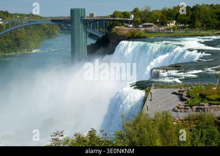 I turisti ammirano le potenti cascate del Niagara dal lato americano, con il Rainbow Bridge e la torre di osservazione sullo sfondo, godendosi la natura Foto Stock
