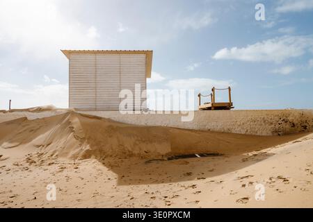 Piccola capanna da spiaggia bianca in legno che sorge accanto ai massicci corsi di sabbia formati dopo le forti tempeste invernali dell'Atlantico sulla spiaggia di Faro in Algarve, Portogallo. Larg Foto Stock