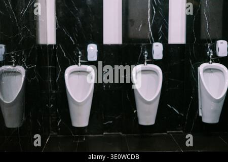 Una fila di orinatoi bianchi puliti e moderni contro un'elegante parete in marmo nero in un bagno pubblico per uomini, che mette in risalto l'igiene e il design contemporaneo. Foto Stock