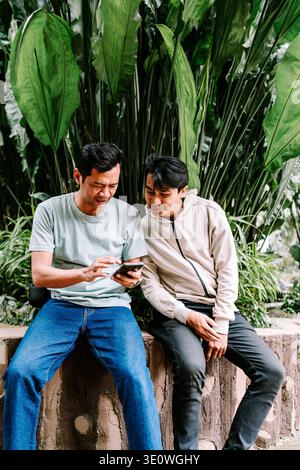 Due giovani uomini asiatici sono seduti all'aperto, concentrati su uno smartphone, condividendo contenuti o navigando insieme in un lussureggiante ambiente tropicale dal verde vivace Foto Stock