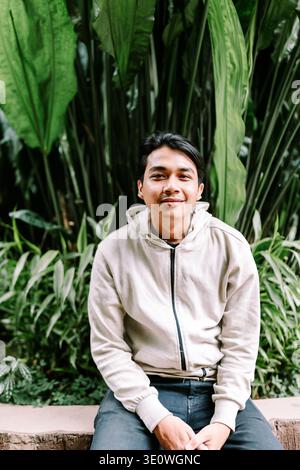 Un giovane asiatico allegro siede comodamente in un vivace giardino verde, sorridendo genuinamente alla macchina fotografica. Indossa una felpa con cappuccio casual, che incarna un modello rilassato Foto Stock