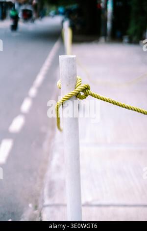 Una corda giallo-verde brillante strettamente legata con un nodo intorno a un palo bianco, adagiata su una strada urbana leggermente sfocata con un marciapiede e una strada in d Foto Stock