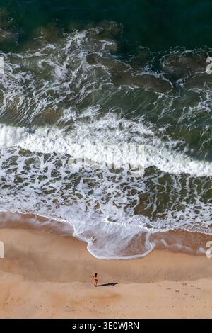 Ripresa aerea ad alto angolo che cattura una donna solitaria in piedi su una spiaggia di sabbia incontaminata mentre le onde turchesi si infrangono sulla riva. La potente schiuma marina bianca Foto Stock
