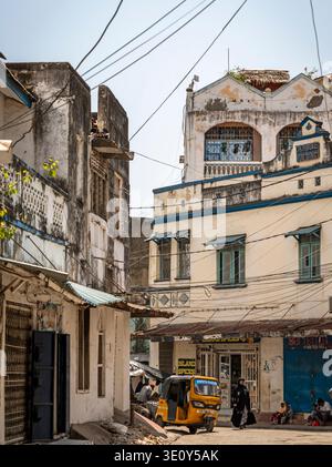 Scena di strada in un'antica città costiera con edifici intemprati, linee elettriche aggrovigliate, piccoli negozi, pedoni e un tuk-tuk giallo sotto il sole Foto Stock