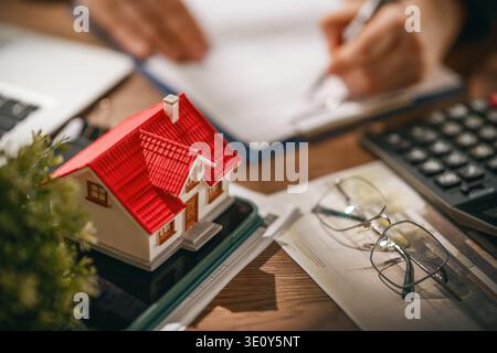 Immagine ravvicinata di una casa in miniatura con un tetto rosso, documenti, calcolatrice, occhiali e una persona che firma documenti, che rappresenta il viaggio emotivo Foto Stock