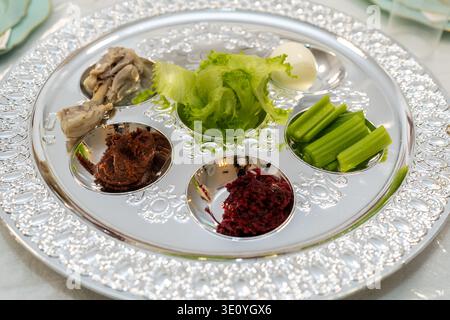 Primo piano del tradizionale seder pasquale con cibi simbolici tra cui lattuga, uovo, sedano, caramello, erbe amare e ossa arrosto su vassoio d'argento decorativo durante le festività ebraiche. Foto Stock