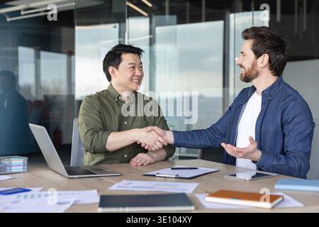 Due partner commerciali diversi stringono la mano, finalizzano un accordo e mostrano partnership, collaborazione e fiducia in un ambiente moderno per riunioni aziendali Foto Stock