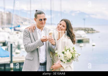 Coppia felice ed elegante che mangia un gelato vicino al porticciolo del lago con montagne sullo sfondo, uomo e donna sorridenti con fiori che si godono momenti romantici durin Foto Stock