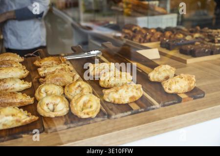 Empanadas marrone dorato e dolci salati disposti su tavole di legno attirano i clienti in una moderna vetrina da forno, che offre piatti freschi pronti da mangiare Foto Stock