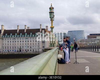 Una coppia maschio e una femmina stanno di fronte a un telefono montato su un treppiede sul Westminster Bridge, Londra. I due stanno filmando se stessi o catturando un ritratto temporizzato a lunga esposizione con l'iconico London Eye del lastminute.com e lo skyline di South Bank come sfondo. Questo tratto del ponte è un luogo di prim'ordine per il "vlogging" e la creazione di contenuti sui social media, nonostante il forte flusso di pendolari pomeridiani e turisti Foto Stock