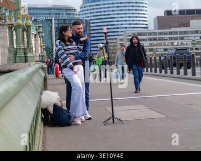 Una coppia maschio e una femmina stanno di fronte a un telefono montato su un treppiede sul Westminster Bridge, Londra. I due stanno filmando se stessi o catturando un ritratto temporizzato a lunga esposizione con l'iconico London Eye del lastminute.com e lo skyline di South Bank come sfondo. Questo tratto del ponte è un luogo di prim'ordine per il "vlogging" e la creazione di contenuti sui social media, nonostante il forte flusso di pendolari pomeridiani e turisti Foto Stock